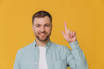 Cute happy unshaven man in mint shirt showing finger up at copy space isolated on yellow background