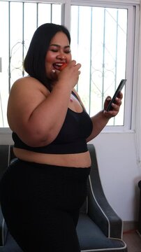 young brunette latina plus size overweight woman wearing sportswear using smartphone and eating apple