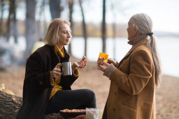 Two woman friends  drinks tea and enjoying a holiday  in autumn outdoor.  portrait of a smiling senior woman with an adult daughter relaxed outdoors 