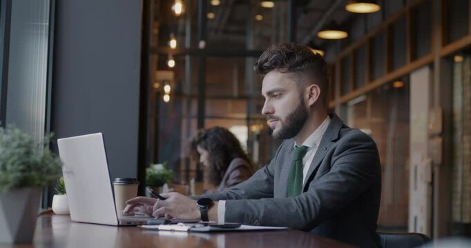 Side view of young businessperson working with computer typing browsing sitting in cafe with group of people. Businessman and modern lifestyle concept.