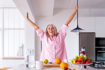 Cheerful mature woman with arms raised standing in kitchen