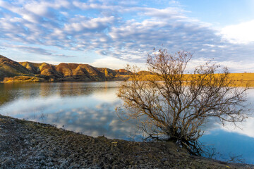 Sunrise at the reservoir. Sunlight on the mountains. Cloudly. Reflections of the mountains and sky. Tree without leaves