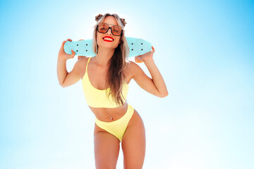 Young beautiful smiling blond woman posing in the street in sunny day. Sexy model in swimwear bathing suit. Positive female with red lips. Holding penny skateboard. Happy and cheerful, blue sky behind
