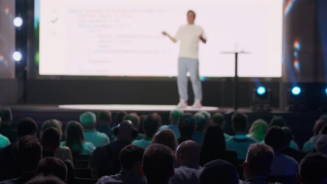 Speaker giving a presentation at a tech conference, engaging with a seated audience. The event takes place in a large venue with bright lights.