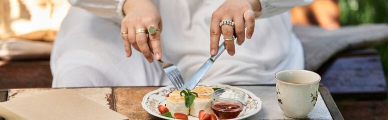 A young woman in a white dress enjoys a summer brunch outdoors, cutting into a savory dish with a knife and fork.