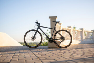 grey road bike is parked against a white wall. The bike has thin tires and is sitting on a concrete surface. The sun is shining brightly in the background