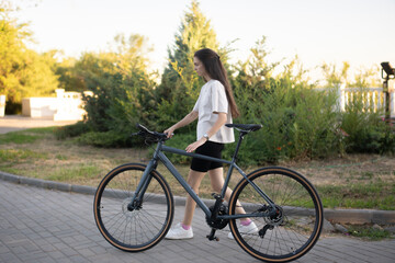 woman in a white shirt walks alongside a black bicycle on a brick path with a bench and a city view in the background, lit by the setting sun