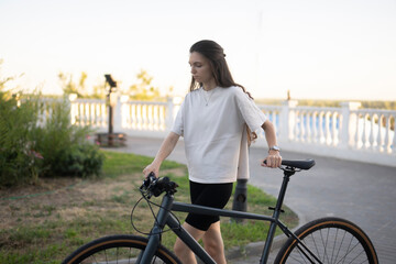 woman in a white shirt walks alongside a black bicycle on a brick path with a bench and a city view...