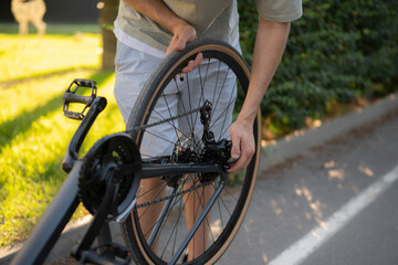Naklejka premium man adjusts the wheel of a bicycle while standing on a paved path. Green grass and trees are in the background