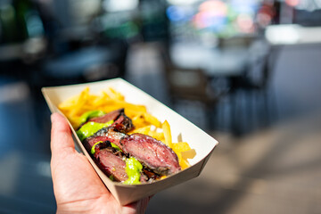 Close-up of a hand holding a paper cone filled with French fries and grilled meat, with a blurred urban background.