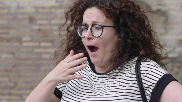 A woman with curly hair yawns dramatically, showing signs of fatigue and boredom. Her open mouth and closed eyes emphasize the intensity of her yawn
