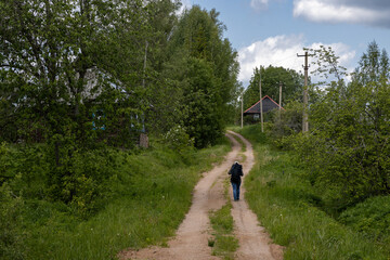 A woman with a backpack walks along a country road towards a village. Rural dirt road. Wooden houses line the village street. Tourist during a hike in the countryside.