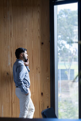 Talking on smartphone, businessman standing by window in modern office, copy space
