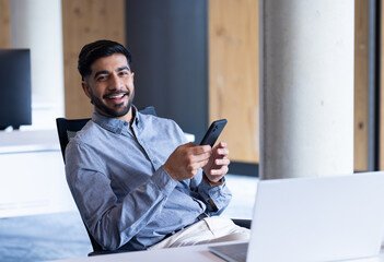 Smiling businessman using smartphone and laptop while sitting in modern office, copy space