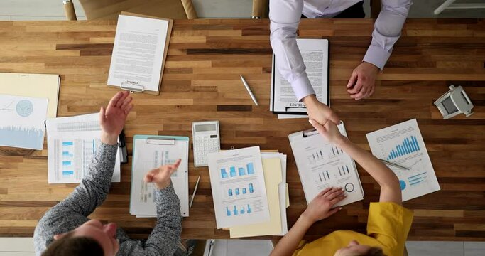 Man claps hands while colleagues doing handshake sitting at wooden table. Happy employees team finish work with investigation papers upper view slow motion