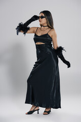 A woman in a black satin dress with feather accents poses against a white backdrop.