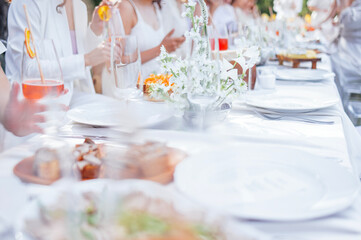 A festive covered table with a tablecloth with cutlery, decorated with flowers. Delicious snacks and glasses with drinks. People in white fancy clothes.