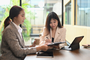 Smiling Asian female colleagues using digital tablet and analyzing financial data at co working...