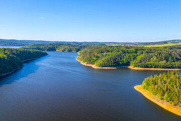 Fototapeta premium Aerial view of Orlik Water Reservoir on the Vltava River in Czechia, surrounded by forests and hills. Calm waters reflect blue sky, creating serene and idyllic landscape.