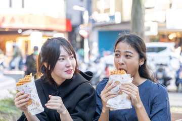Three people - a Taiwanese woman, a Taiwanese man, and an Asian woman - sitting together on a bench in Da’an District, Taipei City, chatting and enjoying a large waffle.