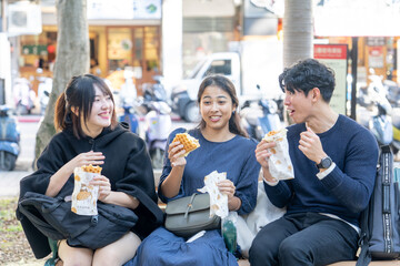 Three people - a Taiwanese woman, a Taiwanese man, and an Asian woman - sitting together on a bench in Da’an District, Taipei City, chatting and enjoying a large waffle.