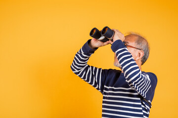 Portrait Asian smiling old man looking camera through binoculars studio shot isolated yellow background, senior man pensioner feeling surprised using binocular