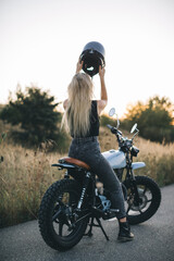 Obraz premium A cute blonde girl puts on a protective helmet while sitting on a motorcycle at sunset.