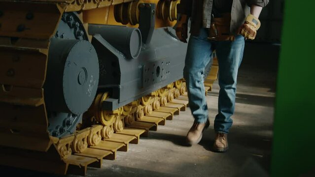 Tilt up shot of male technician in workwear walking along continuous tracked vehicle at industrial machinery plant