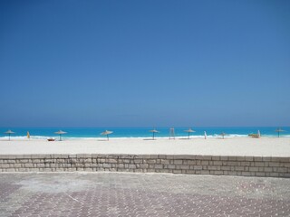 Horizon. Turquoise water of the Mediterranean Sea. White waves near the shore. Beach umbrellas with straw tops line the beach. Snow-white sand. Stone low fence. Outdoor floor tiles. City beach 