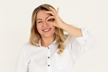 Happy young woman shows ok sign with hand on eye looking through fingers on white background.