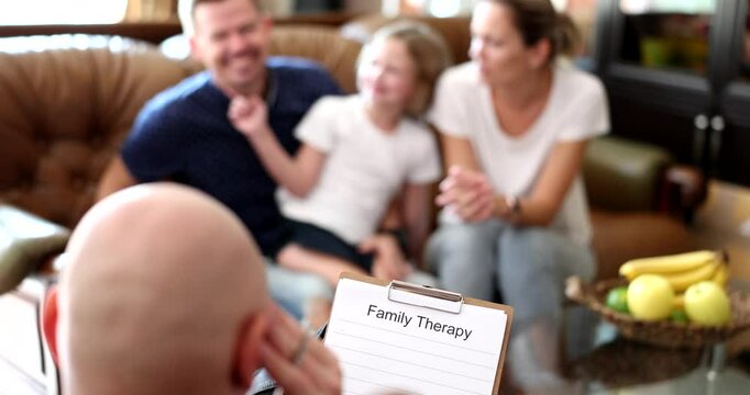 Psychologist holds clipboard ready to make notes on family therapy session. Bald man specialist helps parents understand behavior of little daughter slow motion