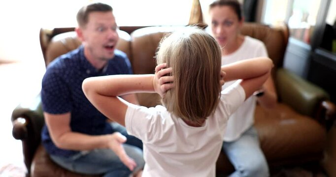 Little girl closes ears with hands refusing to hear angry parents. Emotional father and mother sit on sofa screaming and scolding child slow motion