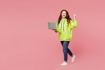 Full body young ginger fun IT woman she wear green hoody casual clothes hold use work on laptop pc computer do winner gesture isolated on plain pastel light pink background studio. Lifestyle concept.