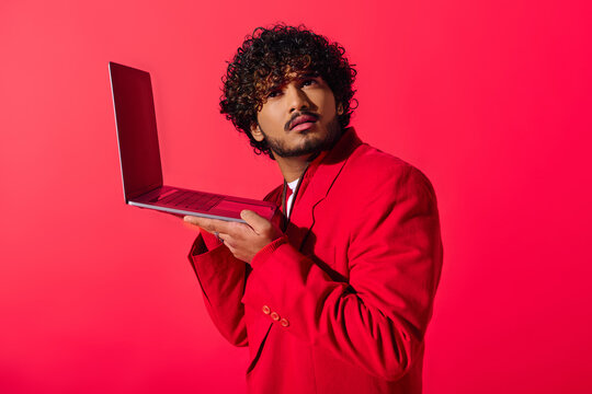 Handsome young Indian man in a vibrant red suit confidently holds a laptop.