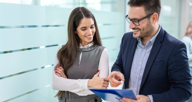 Business woman and man discussing paperwork and looking and pointing at document while walking in office hall.
