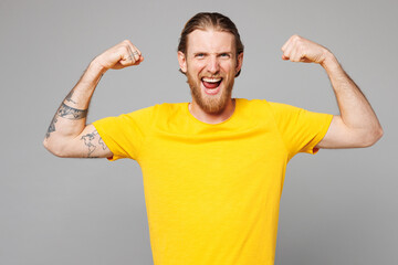 Fototapeta premium Young strong sporty fitness man he wear yellow t-shirt casual clothes showing biceps muscles on hand demonstrating strength power isolated on plain grey background studio portrait. Lifestyle concept.