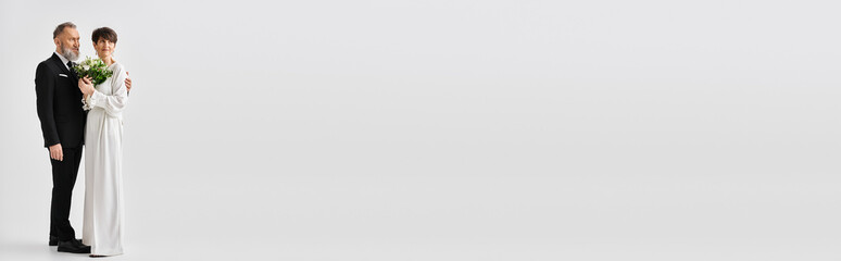 A middle aged bride and groom dressed in wedding gowns celebrating their special day in a studio setting, standing next to each other.