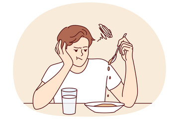 Man suffers from lack of appetite sitting at table in kitchen and does not want to eat breakfast. Depressed guy with unhappy expression melancholy looks at plate with not appetite food during diet