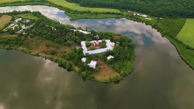 High angle drone footage of Manastirea Caldarusani and it's surrounding lake on a warm summer day. Calm water around a forrest in which a monastery is situated. Aerial shot christian of temple.
