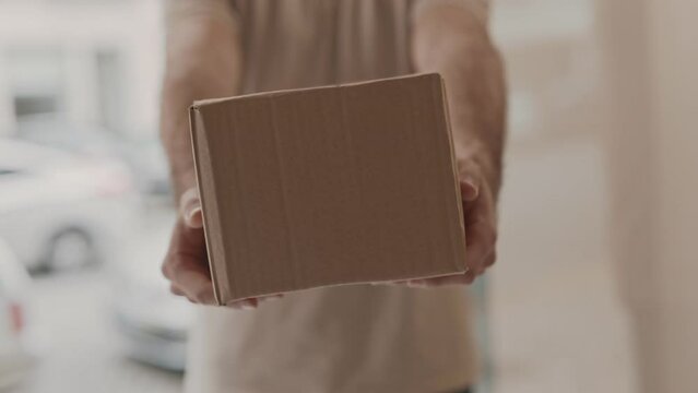 Unrecognizable Close-up shot of a man in casual attire holding a small cardboard box in focus, ready to hand it over in residential area on backdrop, concept of door-to-door delivery.