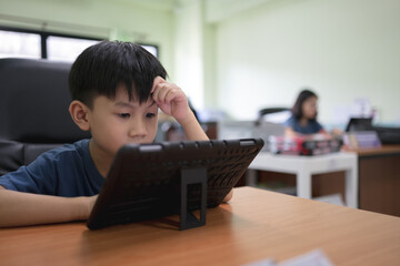 Focused boy using a tablet at a desk in a classroom with his mother working in the background.