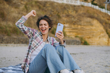 Young happy cool woman wear shirt casual clothes hold in hand use mobile cell phone do winner gesture sit rest on sea ocean sand shore beach outdoor seaside in summer day free time. Lifestyle concept.