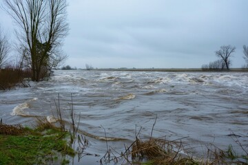 A vast flooded river overflows its serene banks in a rural area, with ferocious waters and submerged fields under a cloudy and somber sky, highlighting nature’s raw power