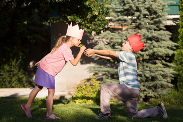 a boy and a girl in paper crowns play in a summer garden. The boy stands on one knee and gives the girl a flower.