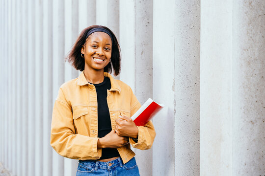 Young woman wearing a yellow jacket and a black shirt is holding a red book. She is smiling and she is happy - Powered by Adobe