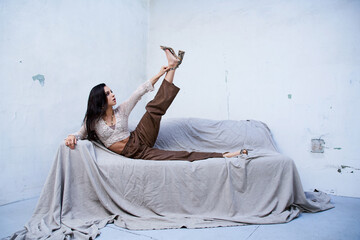 A young woman in a white knitted top and brown pants poses on a gray studio couch.