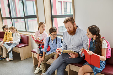 Fototapeta premium A man, the teacher, sits before a group of kids in a bright and lively classroom, animatedly reading a book to captivate their imaginations.