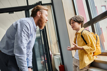 A man stands beside a little boy in front of a window, engaging in a thoughtful conversation while...