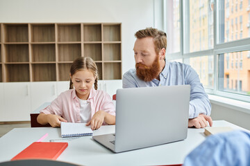 A man and a little girl sit attentively in front of a laptop, engaging in educational content during a lively classroom session.