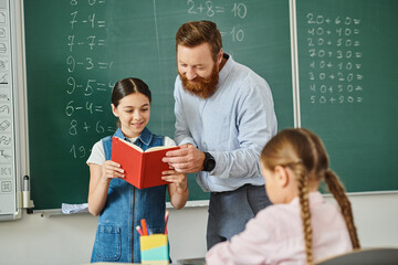 A man teacher sharing a book with a young girl in a colorful classroom filled with students eagerly...